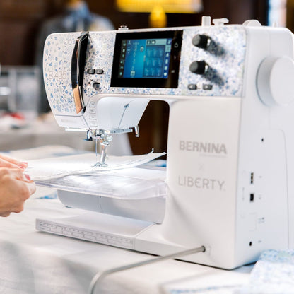 A white Bernina Liberty sewing machine with a digital screen and pattern arm, shown in a real-world setting with a person's hands guiding fabric under the needle.