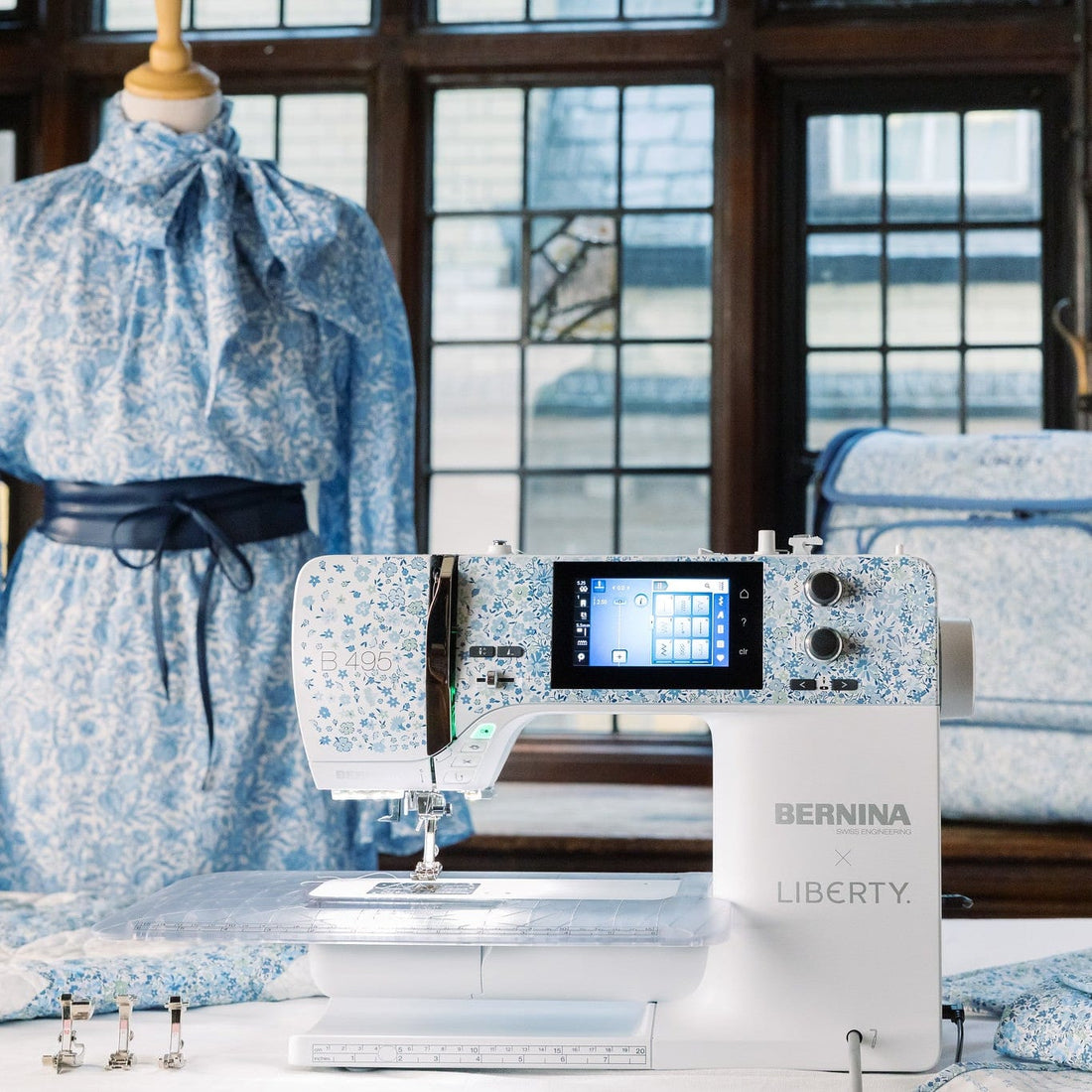 A BERNINA LIBERTY sewing machine in white with a blue floral pattern, displayed in a room with a matching blue floral dress on a mannequin and a blue floral quilt in the background.
