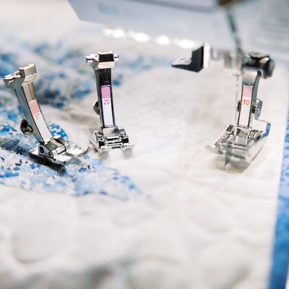 Three metallic sewing machine pressers stitching a white quilted fabric with blue patterned edges under bright studio lighting.