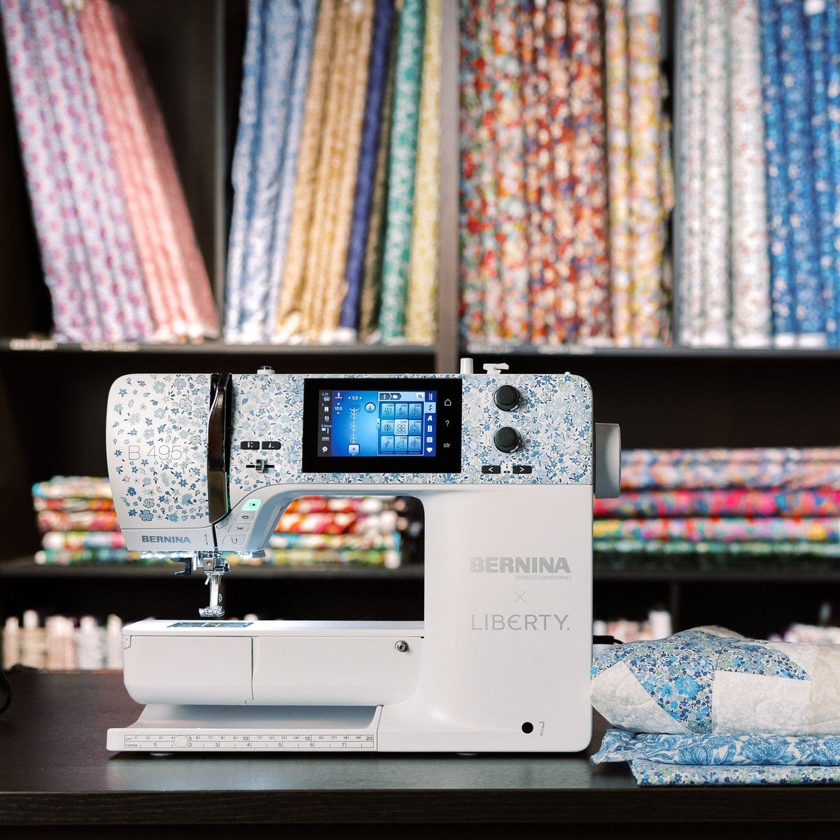 A BERNINA Liberty sewing machine in white with blue floral patterns on the upper arm and control panel, set on a dark table in front of shelves filled with colorful fabrics. A blue patterned garment is draped on a mannequin to the right.