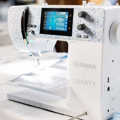 A BERNINA Liberty sewing machine in white with blue floral-patterned accents, shown in a softly lit indoor setting with a blurred lamp and mannequin in the background.