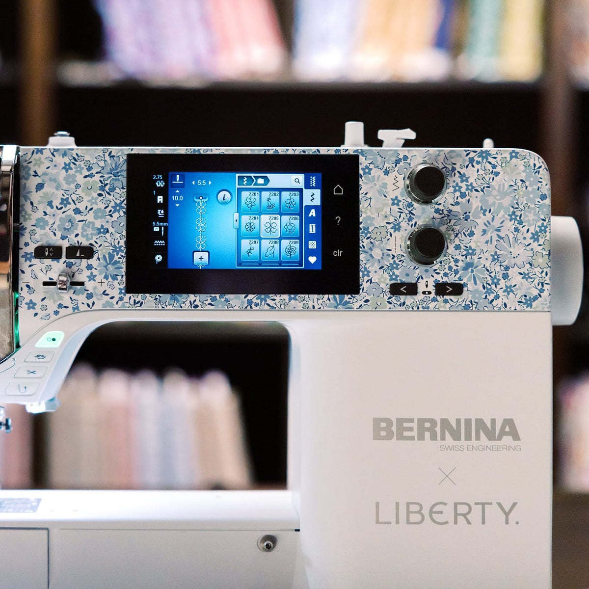 A BERNINA x LIBERTY B-195 sewing machine with a white body and a blue floral patterned upper cover, shown in a retail or showroom setting with blurred shelves in the background.