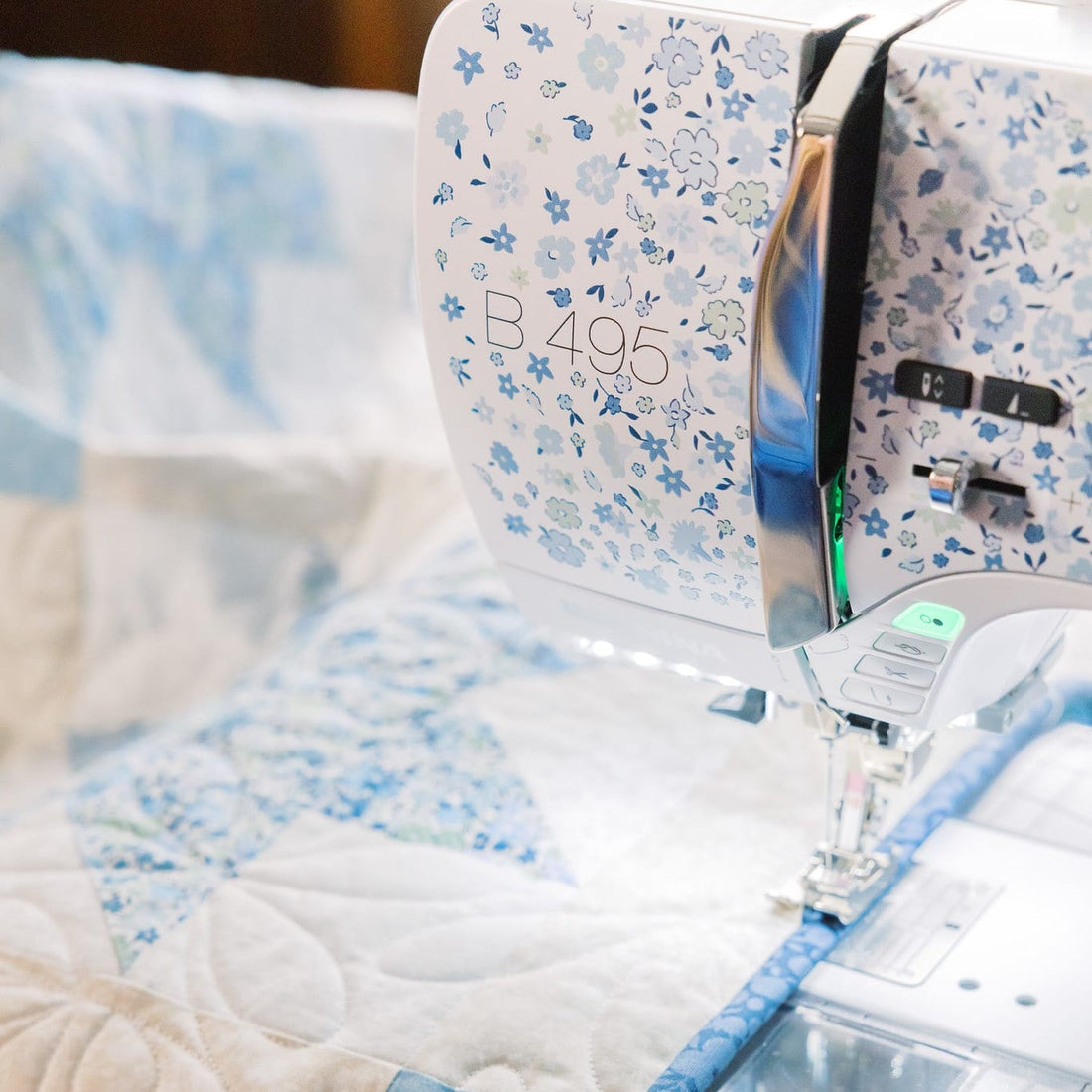 A close-up of a white and blue floral sewing machine stitching a patchwork quilt with light blue and cream fabric sections.