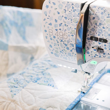 A close-up of a white and blue floral sewing machine stitching a patchwork quilt with light blue and cream fabric sections.