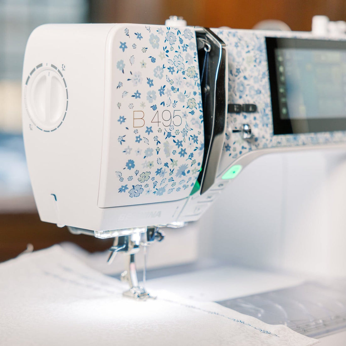 A close-up of a white and blue floral-patterned sewing machine, model B495, stitching white fabric. The background is a softly blurred indoor setting with warm wooden tones.