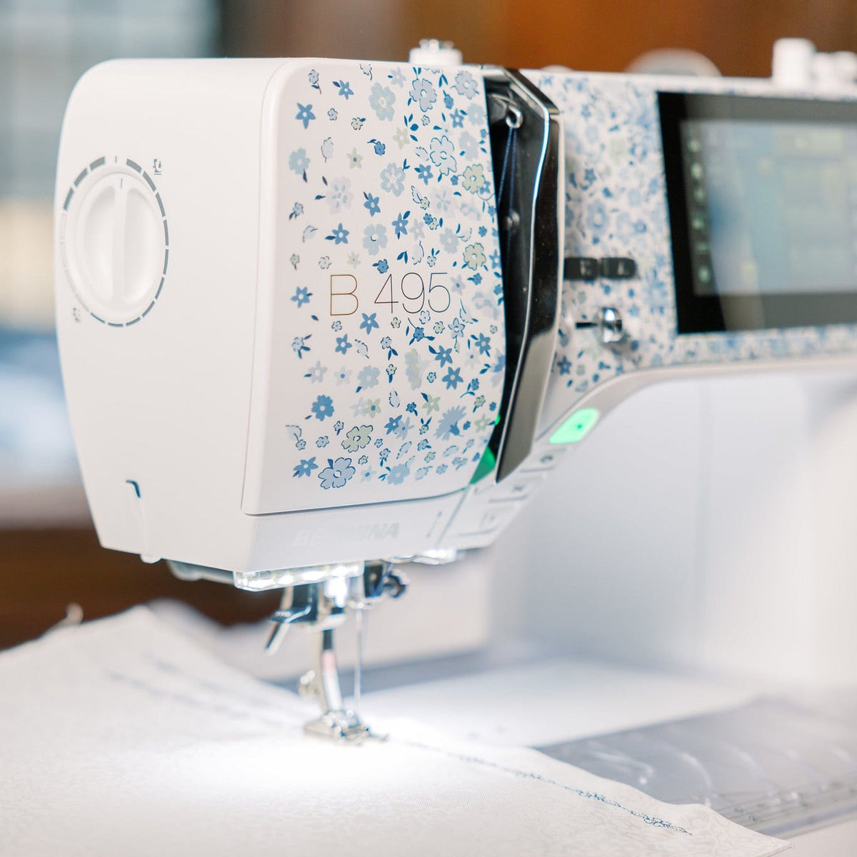 A close-up of a white and blue floral-patterned sewing machine, model B495, stitching white fabric. The background is a softly blurred indoor setting with warm wooden tones.