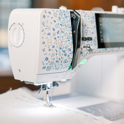 A close-up of a white and blue floral-patterned sewing machine, model B495, stitching white fabric. The background is a softly blurred indoor setting with warm wooden tones.
