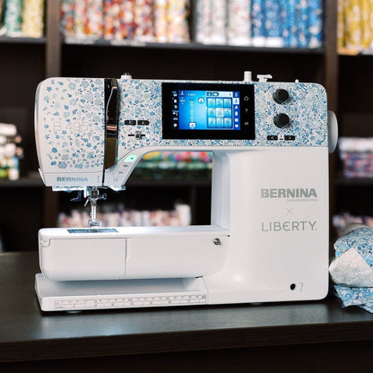 A Bernina Liberty sewing machine sits on a dark table in a sewing room, with a blue and white patterned dress displayed on a mannequin to the right and shelves of colorful fabric in the background.