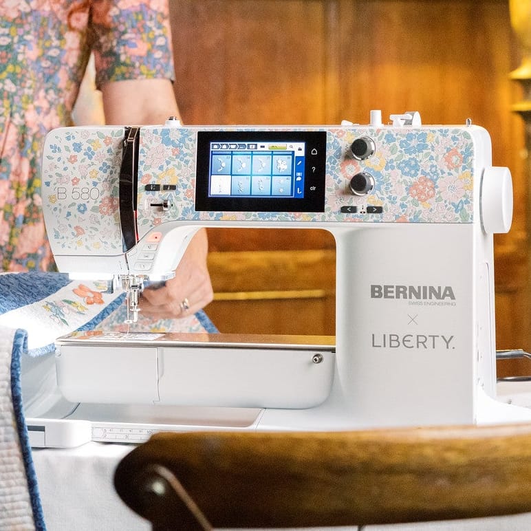 A woman with red hair stands beside a BERNINA LIBERTY sewing machine in a warmly lit room with wood paneling and a lamp, working with a quilt on a table.