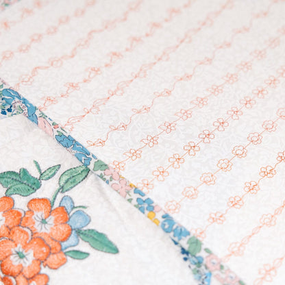 Close-up of a white quilted fabric with delicate peach embroidered floral patterns and a colorful floral border featuring orange flowers, green leaves, and blue accents.
