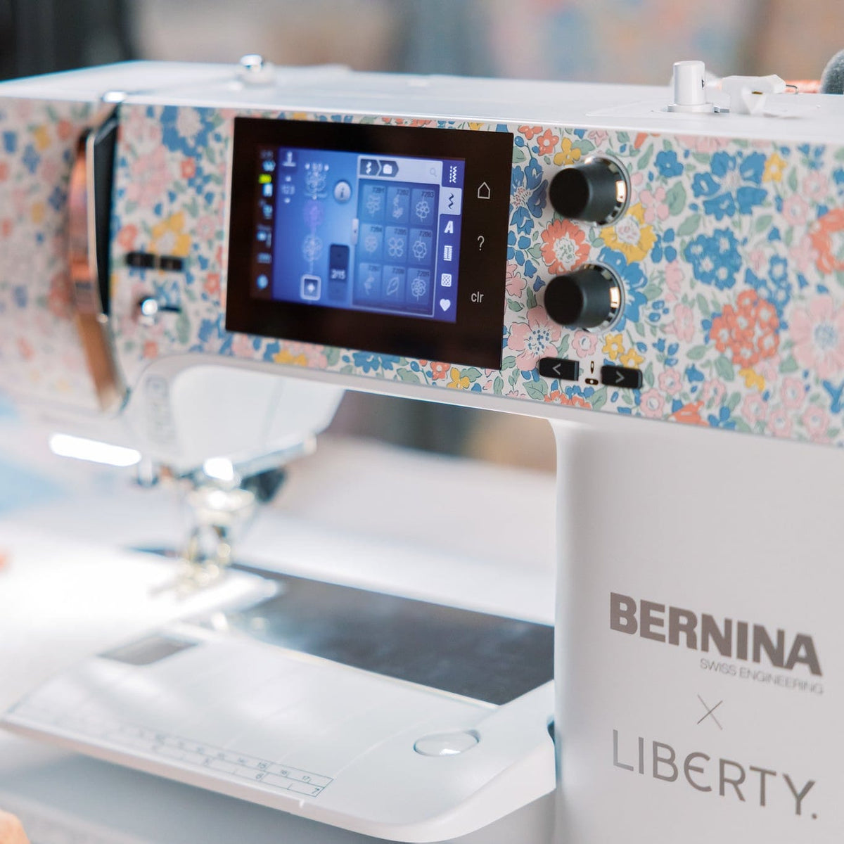 A close-up of a BERNINA x LIBERTY sewing machine with a white body and a colorful floral pattern on the upper panel, with a digital display screen and control knobs. A person's hands are visible guiding fabric under the needle in the foreground.