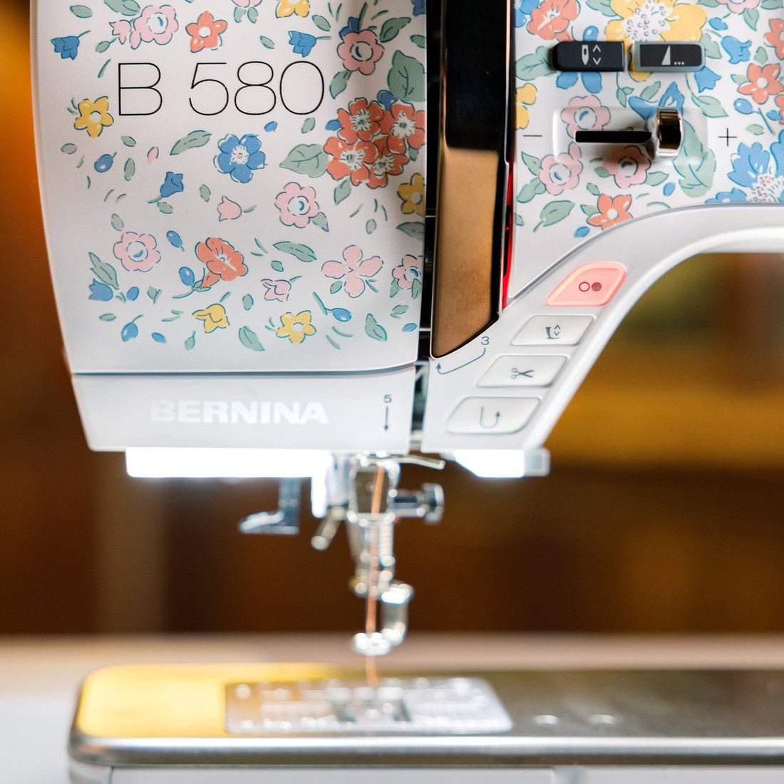 Close-up of a BERNINA B 580 sewing machine with a white body decorated in a colorful floral pattern, set against a warm, blurred wooden background.