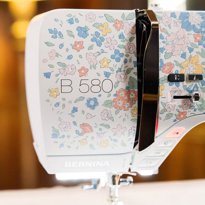 A close-up of a BERNINA B 580 sewing machine with a white body decorated in a pastel floral pattern, set against a warm, blurred indoor background.