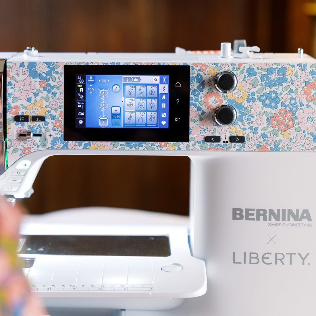 A close-up of a BERNINA B580q embroidery machine with a white body, floral patterned top panel, and a digital display screen, with a person's shoulder visible in the blurred foreground on the left.