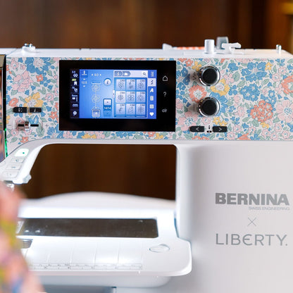 A close-up of a BERNINA B580q embroidery machine with a white body, floral patterned top panel, and a digital display screen, with a person's shoulder visible in the blurred foreground on the left.