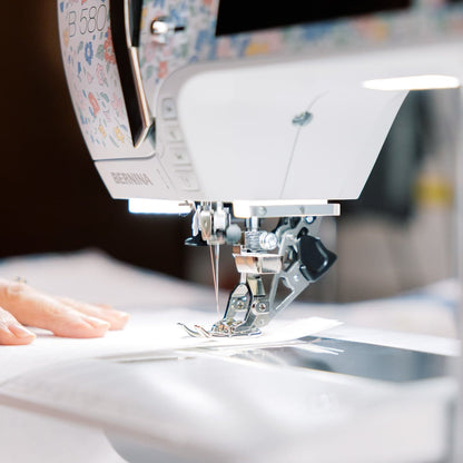 A close-up of a white sewing machine with a floral-patterned upper section, shown in use with a person's hands guiding fabric under the needle.