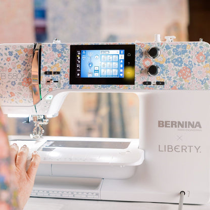 A close-up of a BERNINA x LIBERTY sewing machine with a floral-patterned top cover and a white body, shown in a real-world setting with a person's hand and shoulder visible on the left.