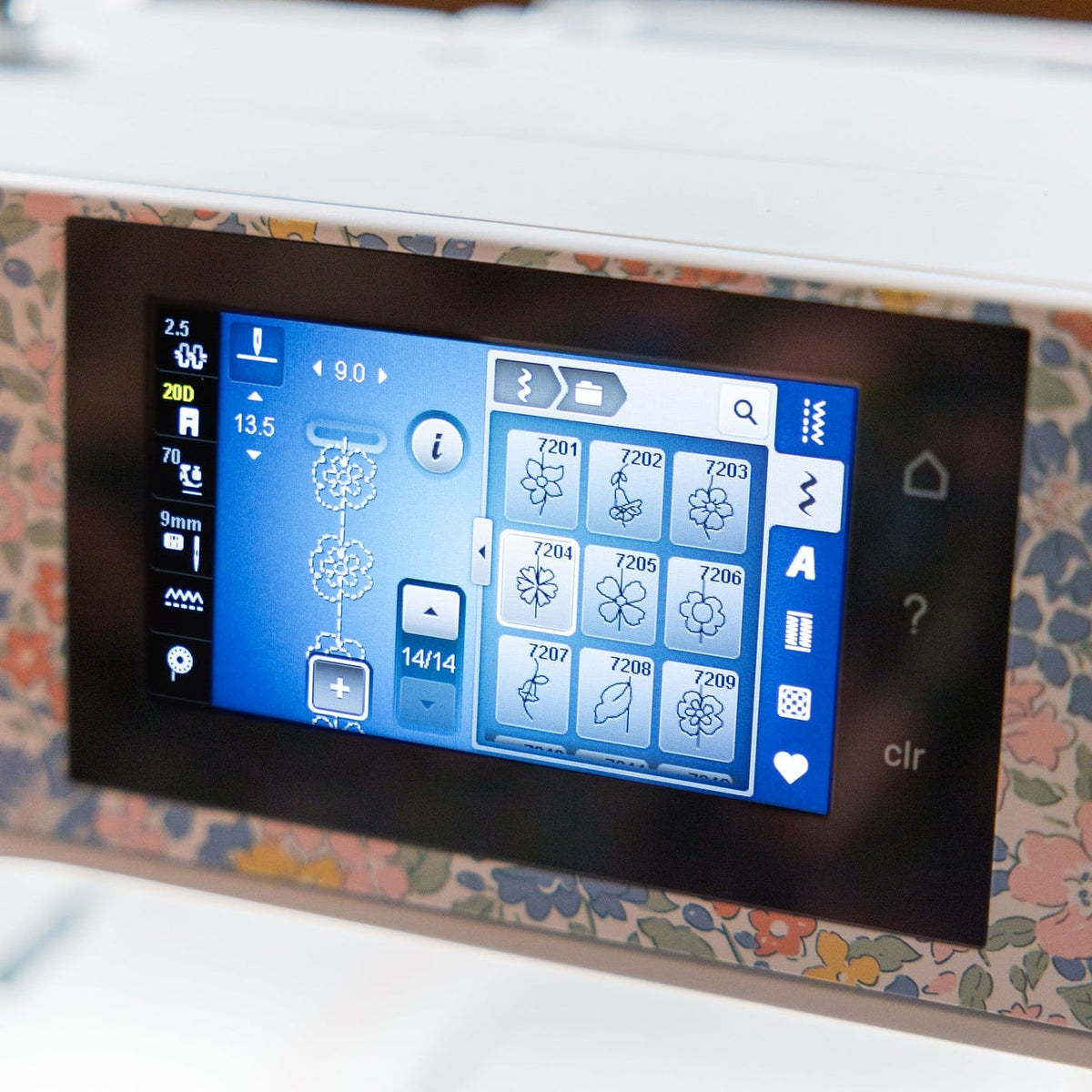 Close-up of a sewing machine touchscreen display showing interface icons, with a floral-patterned front cover and black control knobs visible.