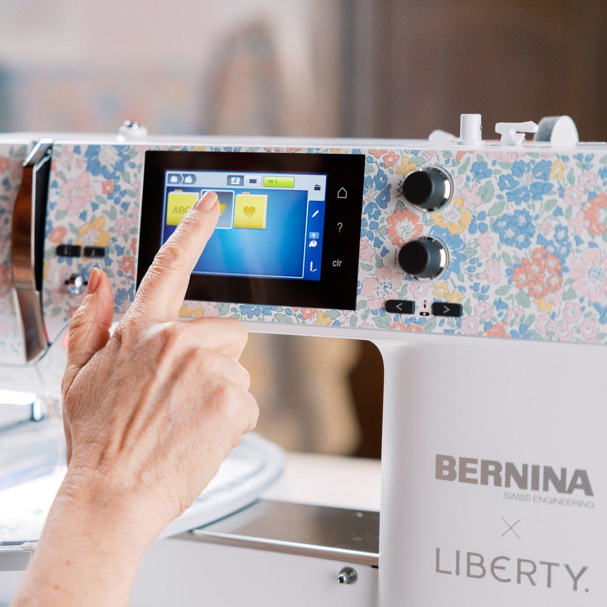 A close-up of a Bernina Liberty sewing machine with a floral patterned top panel and a touchscreen display being touched by a person's hand.