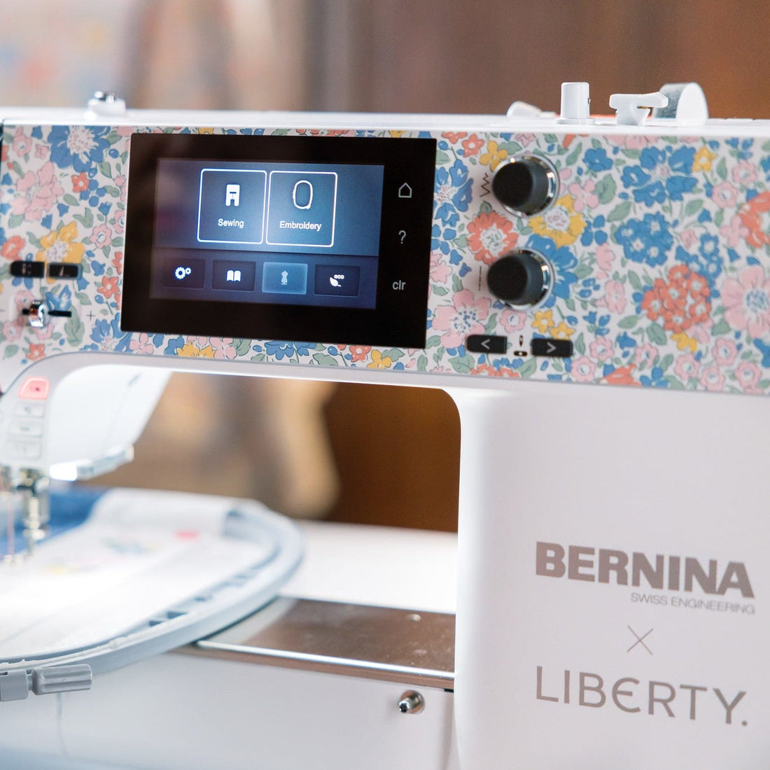 A close-up of a BERNINA Liberty embroidery machine with a white body, a colorful floral patterned top panel, and a digital display screen, with embroidery thread visible in the needle area.