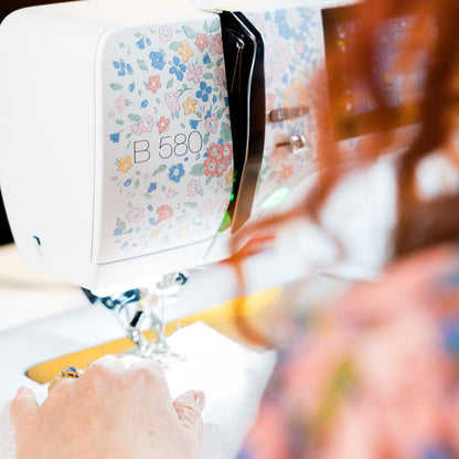 A close-up lifestyle shot of a white B&Q 580 sewing machine with a pastel floral pattern on the front panel, being operated by a person with red hair in a softly blurred indoor setting.