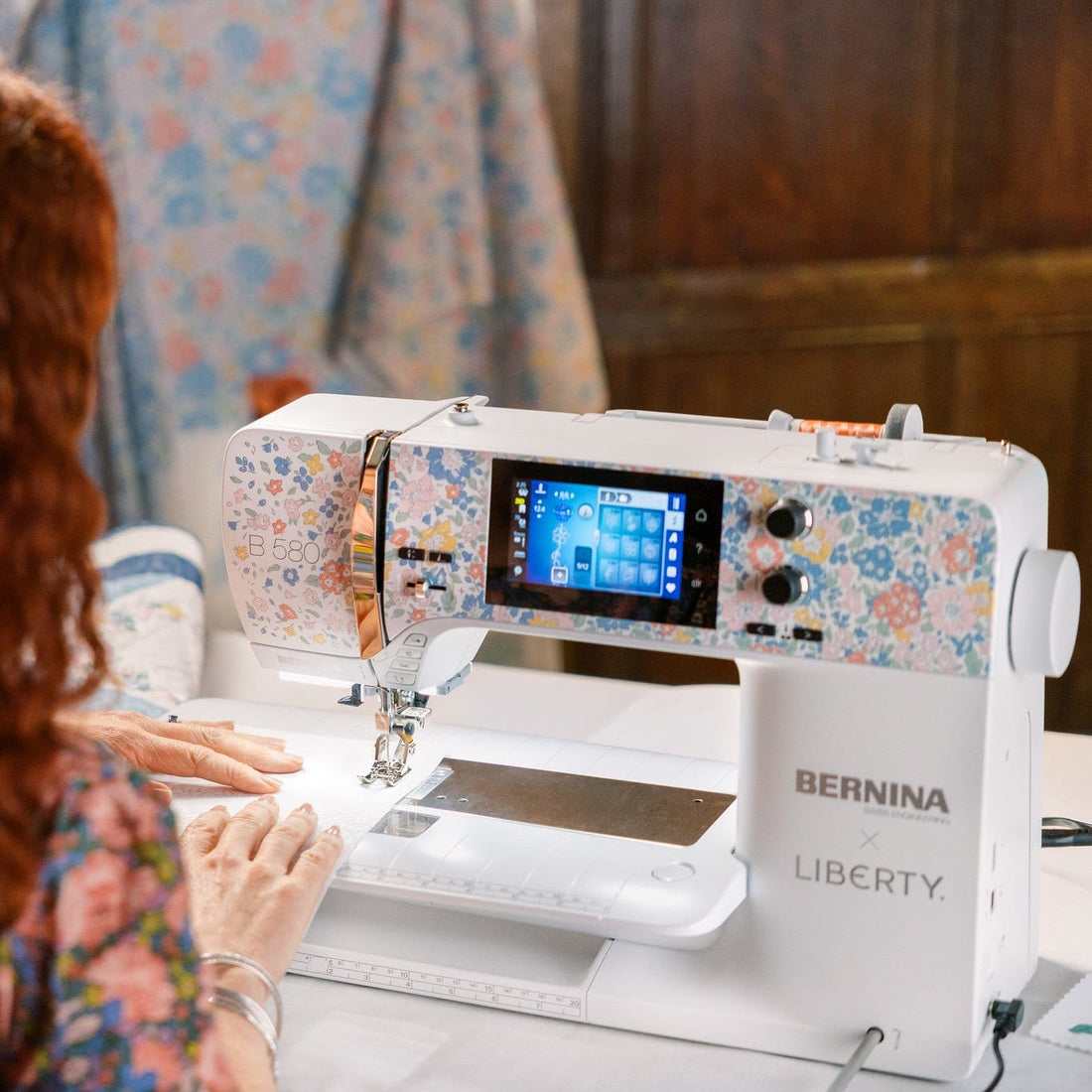 A woman with long curly red hair uses a BERNINA LIBERTY sewing machine with a floral patterned cover, viewed from behind in a home or studio setting.