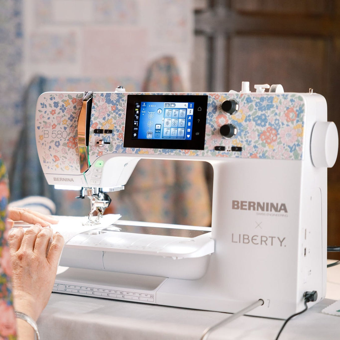 A woman in a floral dress uses a BERNINA x LIBERTY sewing machine with a colorful floral patterned body and a digital screen, in a softly lit indoor setting.
