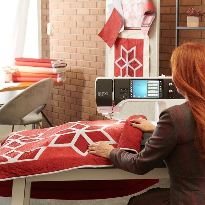 A woman with long red hair sews a red and white patterned quilt on a white sewing machine in a bright craft room with a brick wall background.