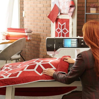 A woman with long red hair sews a red and white patterned quilt on a white sewing machine in a bright craft room with a brick wall background.