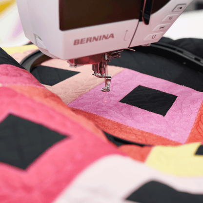A close-up of a BERNINA sewing machine stitching a colorful quilt with pink, yellow, orange, and black geometric blocks.