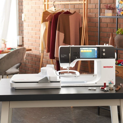 A white and black Bernina sewing machine sits on a dark table in a styled room with a brick wall, clothing rack, and shelving in the background.