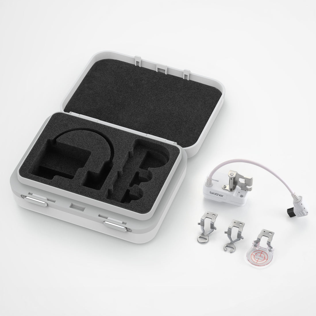 An open light gray hard carrying case with black foam inserts, shown on a white background alongside a small white electronic module with a cable, three metal clips, and a round clear accessory.
