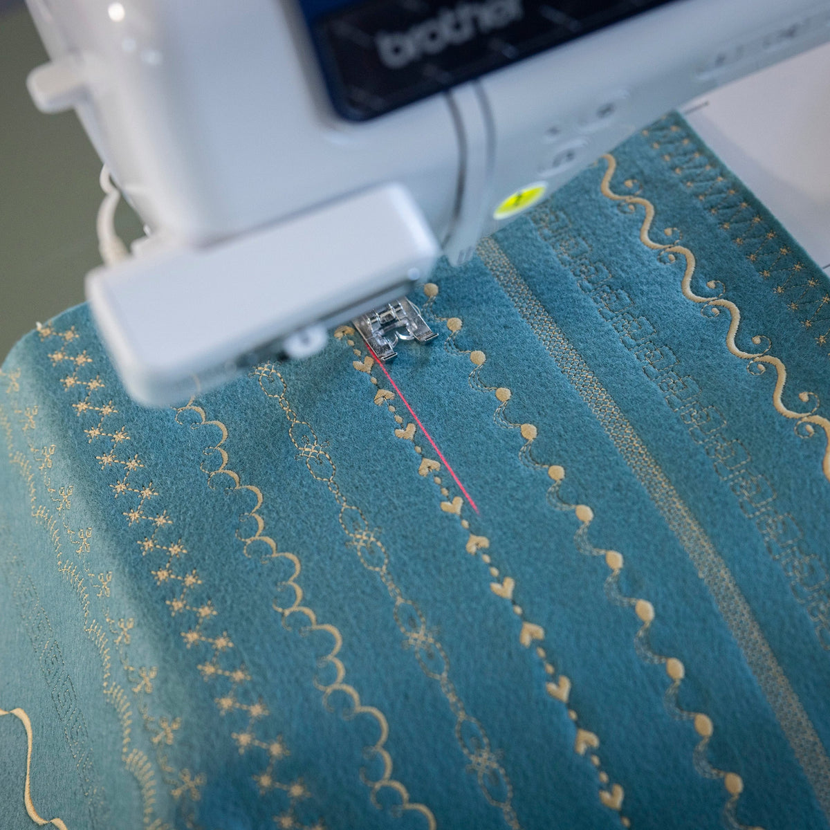 A close-up of a Brother embroidery machine stitching a teal fabric with gold embroidered patterns.