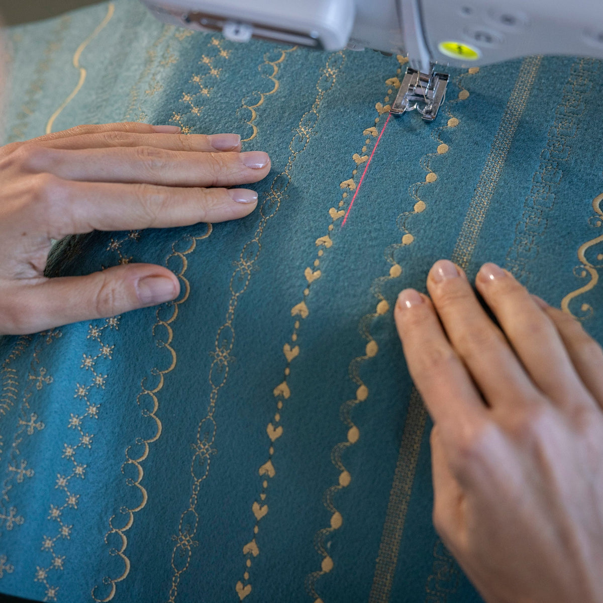 Close-up of a person's hands guiding teal fabric with gold embroidered patterns through a white sewing machine.