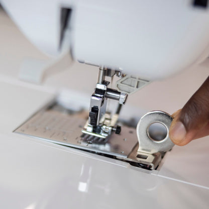 A close-up of a person's hand adjusting a silver metal tension dial on the presser foot area of a white sewing machine.