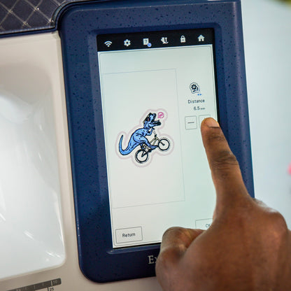 A close-up of a person's finger touching the screen of an embroidery machine, with a blue and white embroidery design visible on the touchscreen. A stack of colorful fabric samples sits on the table in the blurred background.