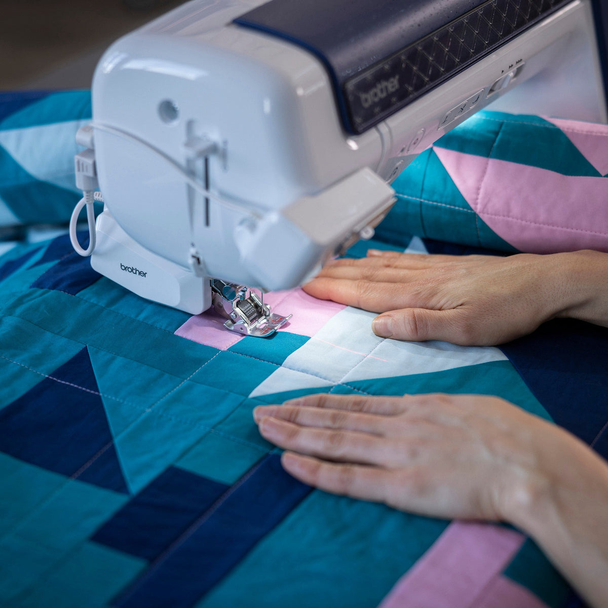 A close-up of a Brother sewing machine stitching a geometric quilt with teal, pink, and white blocks, with a person's hands guiding the fabric under the needle.