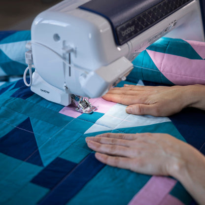 A close-up of a Brother sewing machine stitching a geometric quilt with teal, pink, and white blocks, with a person's hands guiding the fabric under the needle.