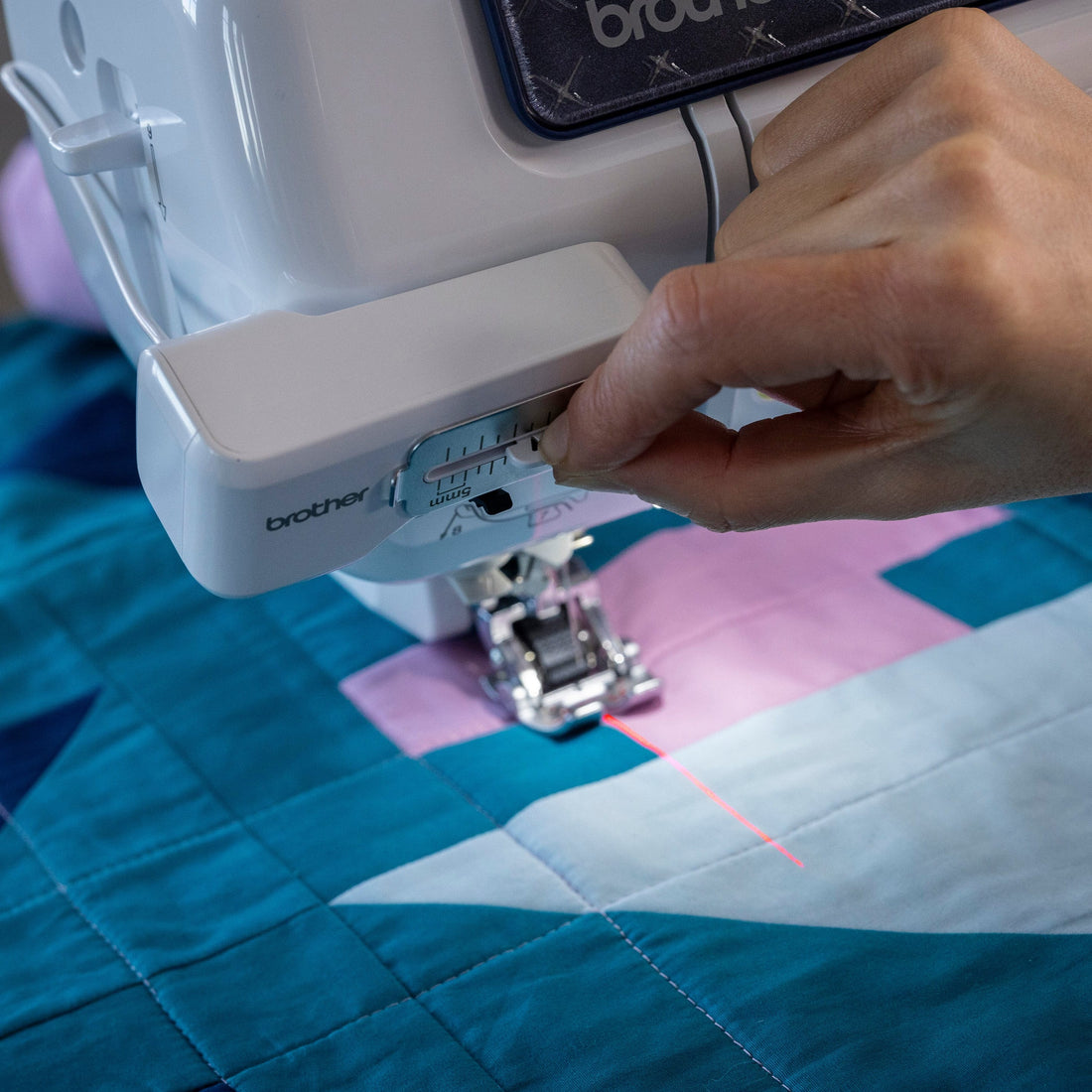 A close-up of a Brother sewing machine being used to quilt a geometric patchwork fabric with blue, pink, and white sections. A person's hand is adjusting the thread tension dial on the white machine body.