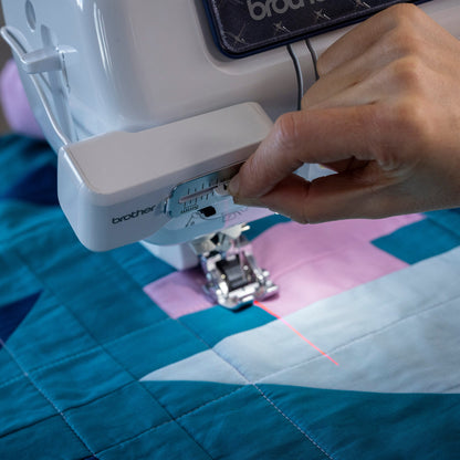 A close-up of a Brother sewing machine being used to quilt a geometric patchwork fabric with blue, pink, and white sections. A person's hand is adjusting the thread tension dial on the white machine body.