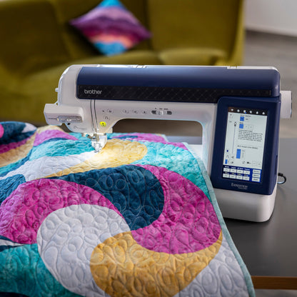 A Brother sewing machine with a digital touchscreen display sits on a dark table, stitching a colorful quilt with pink, teal, yellow, and white patterns.