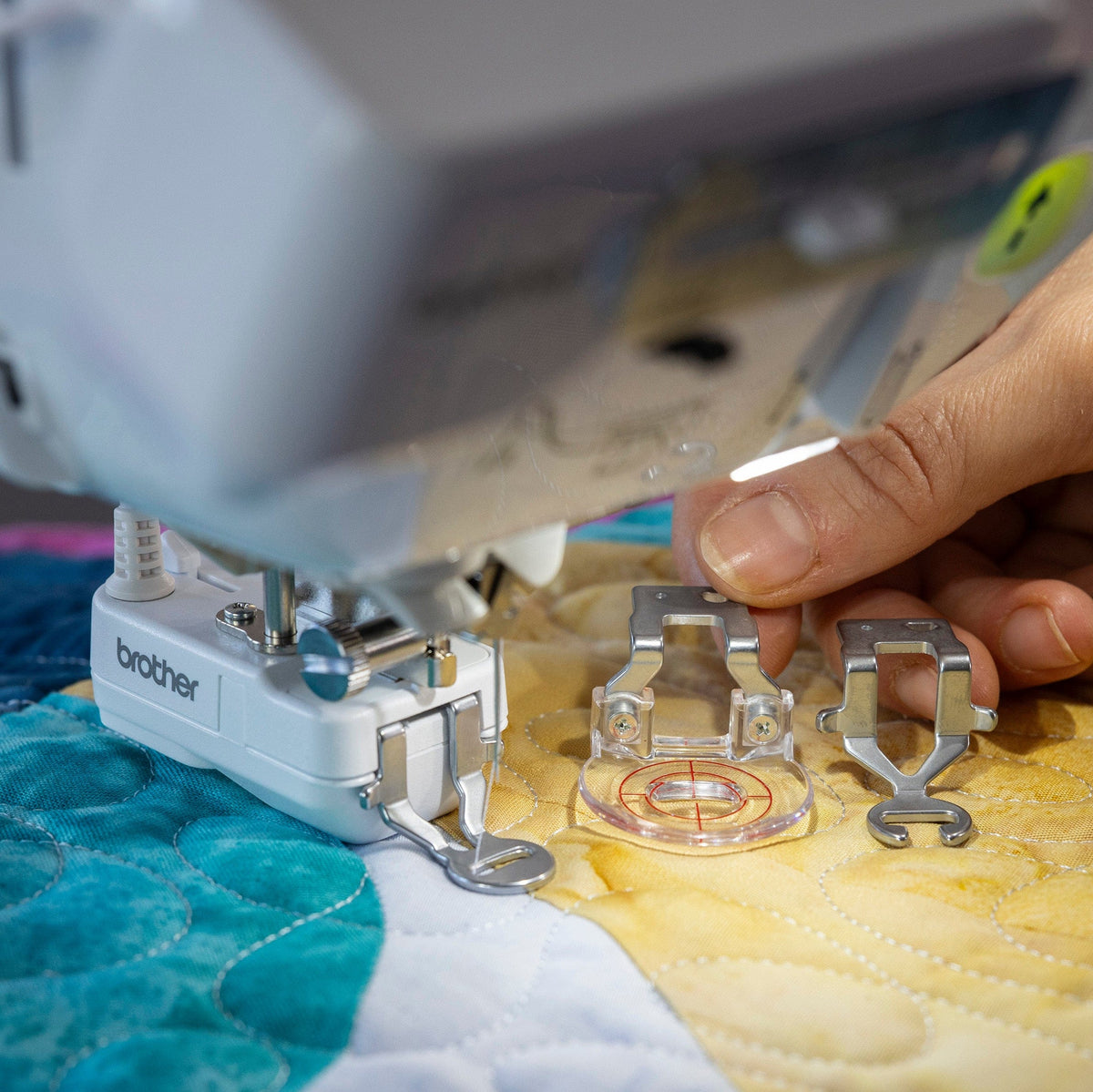 A close-up of a Brother sewing machine in use, with a hand adjusting clear presser foot accessories over a colorful quilted fabric.