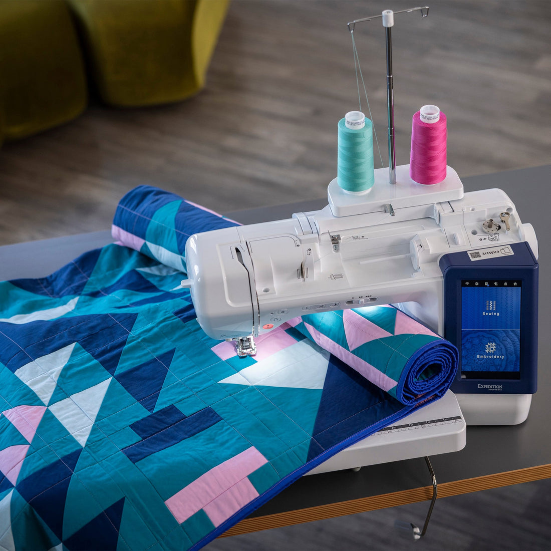 A white sewing machine with a blue digital display screen sits on a gray table, sewing a geometric patterned quilt in shades of blue, teal, and pink. Two spools of thread, one light blue and one pink, are mounted on top of the machine.
