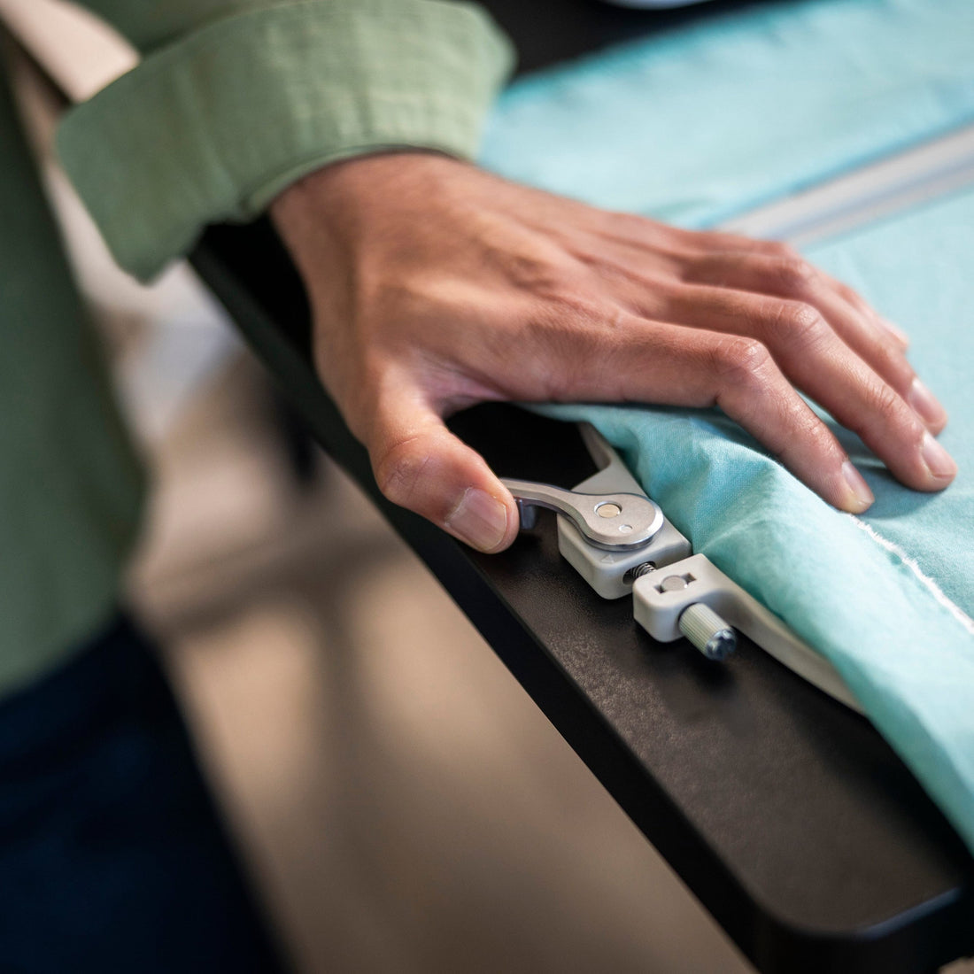 A close-up of a person's hand operating a white and silver electronic fabric measurer on a light blue garment laid out on a black table.