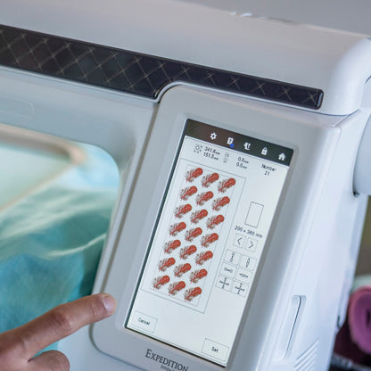 A close-up of a Brother embroidery machine with a touchscreen interface, showing a finger pointing at the screen. The machine is white with a black top panel, and light blue fabric is visible in the embroidery hoop.
