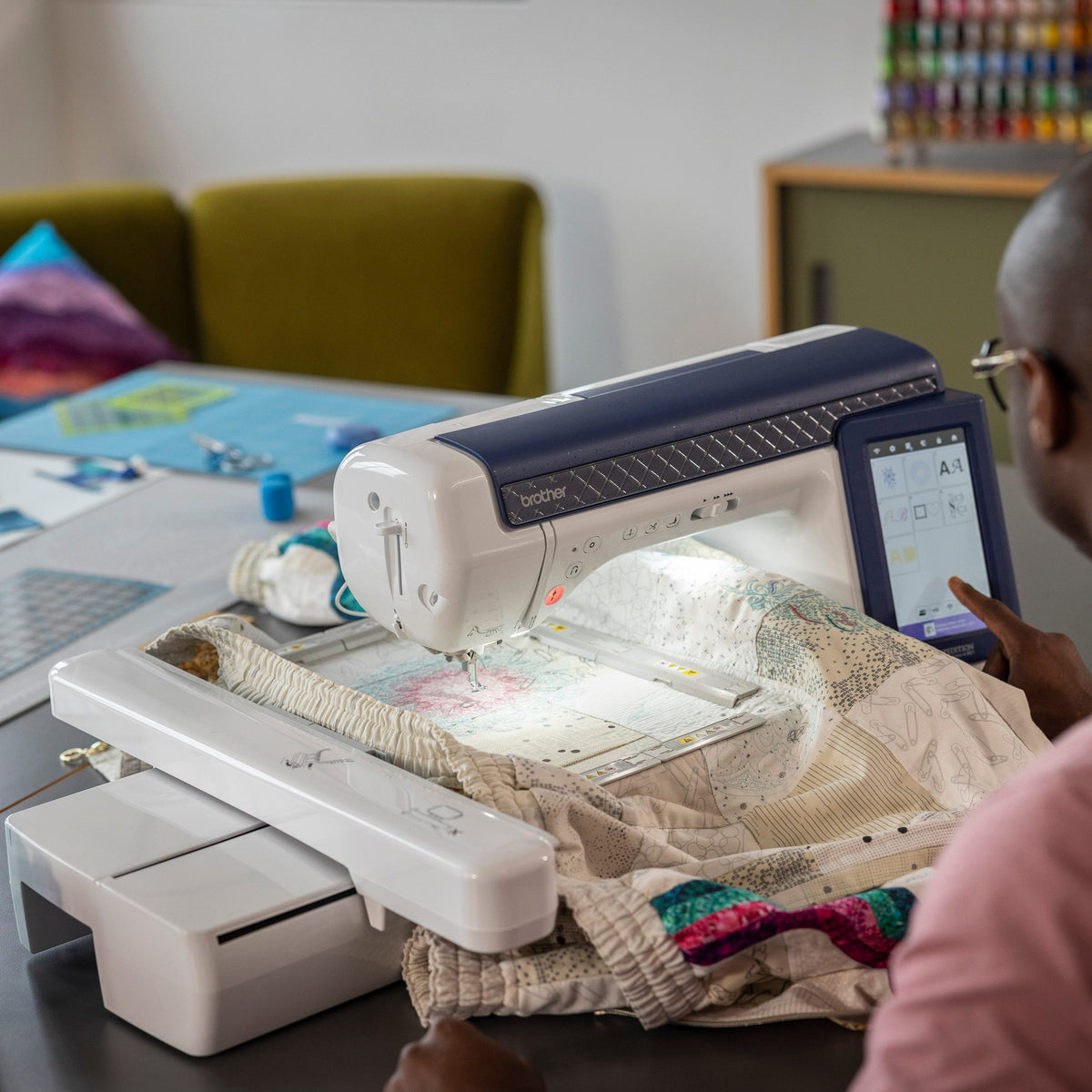 A person in a pink shirt uses a white and navy computerized sewing machine to quilt a cream-colored fabric piece on a dark table, with sewing supplies and green chairs in the background.