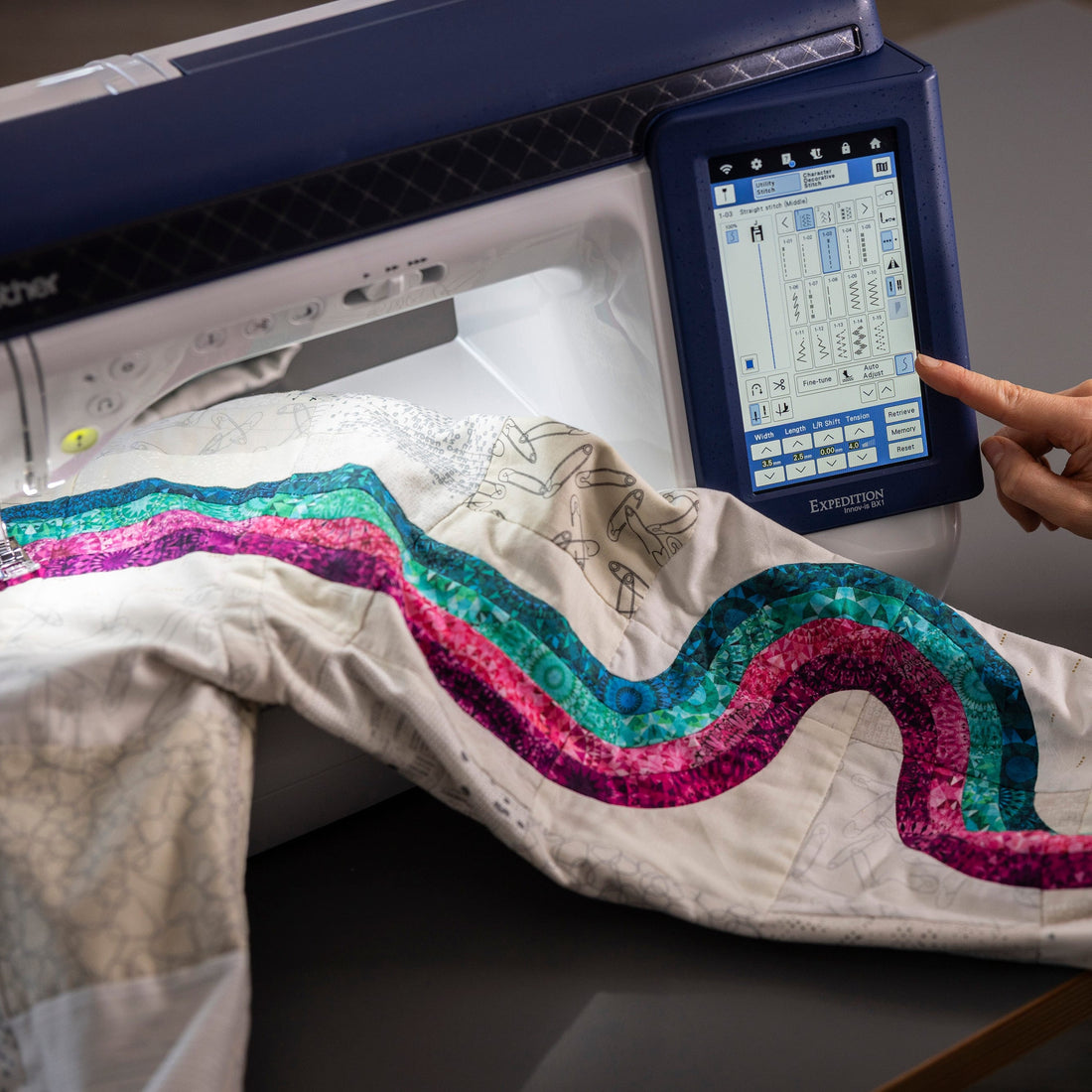 A Brother embroidery machine with a person's hand touching the touchscreen interface, embroidering a wavy multicolor sequin pattern onto a white fabric sleeve.