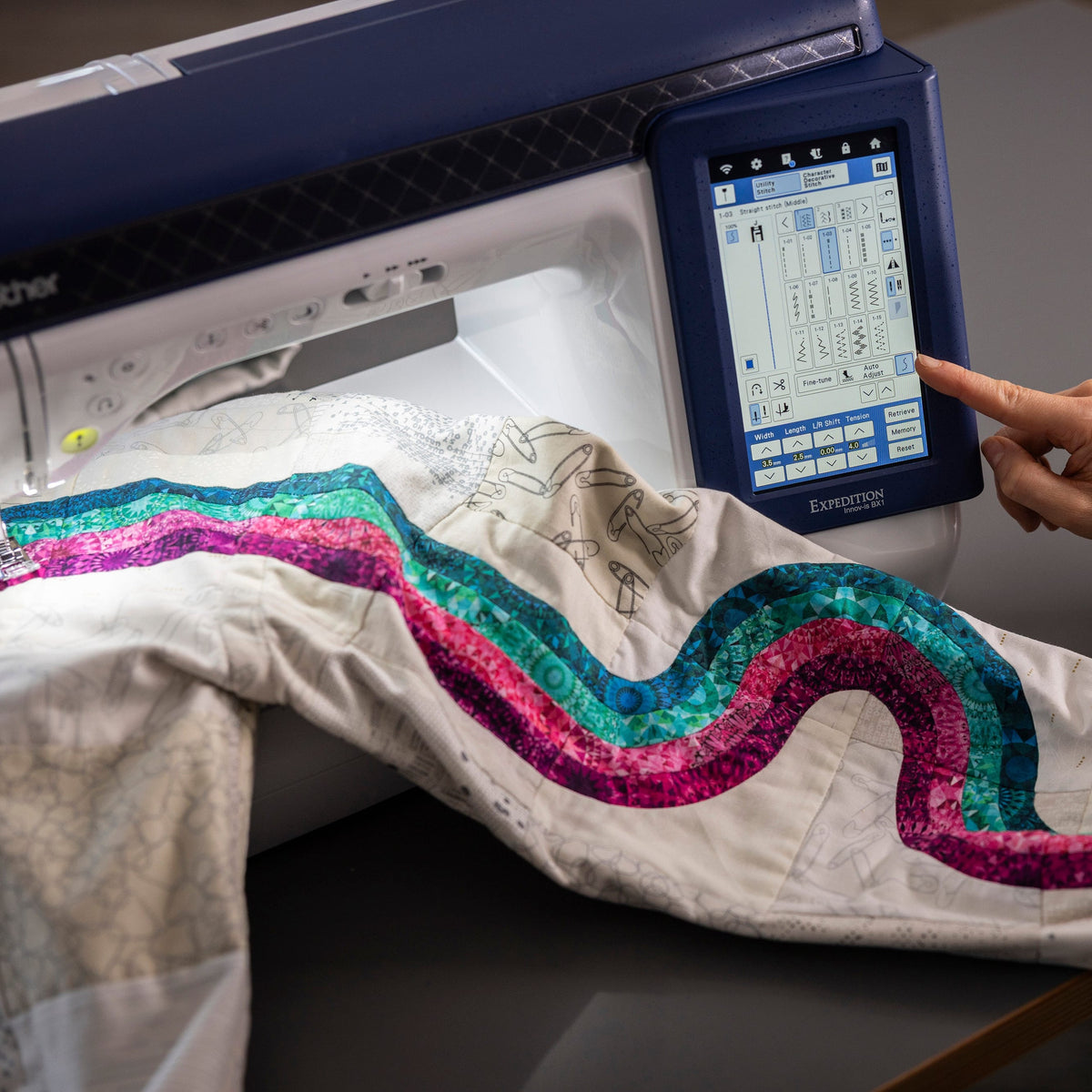 A Brother embroidery machine with a person's hand touching the touchscreen interface, embroidering a wavy multicolor sequin pattern onto a white fabric sleeve.