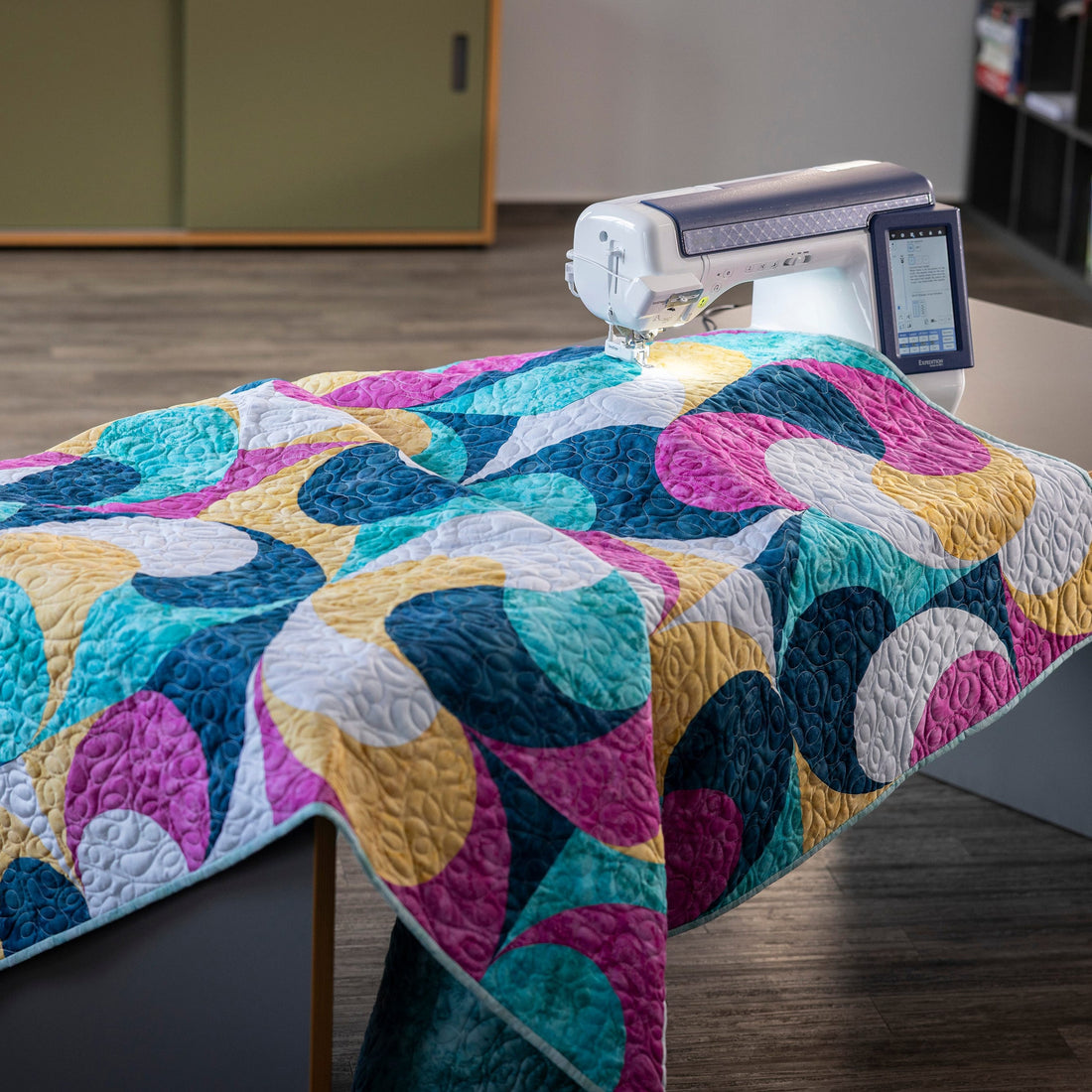 A colorful quilted patchwork blanket with abstract curved shapes in teal, magenta, yellow, and white is draped over a dark table in a room, with a sewing machine on top.