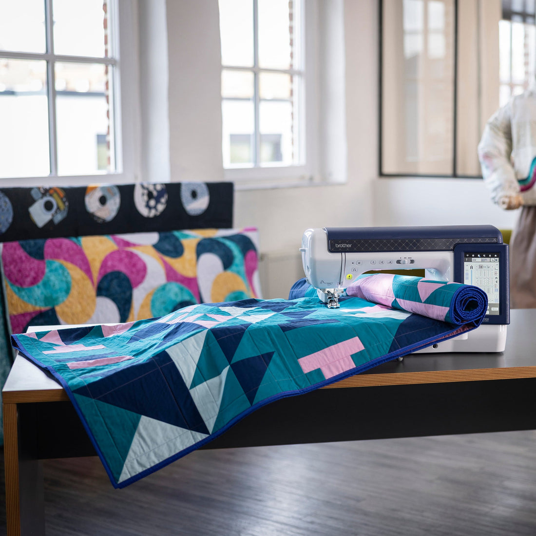 A colorful geometric quilt in shades of teal, navy, pink, and white is draped over a sewing machine on a wooden table in a bright, modern room. A mannequin wearing a patterned sweatshirt stands in the background.